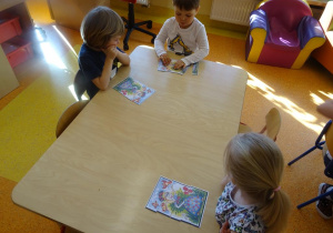 Troje dzieci siedzi przy stole i przygląda się obrazkowi łąki.